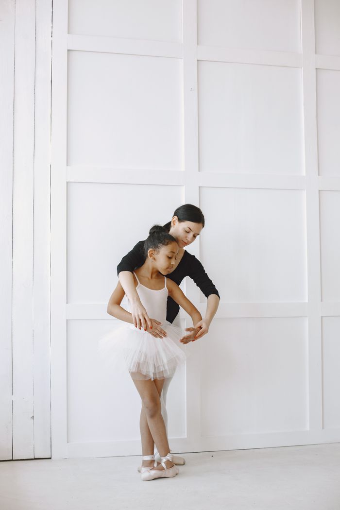 Ballet teacher guides young girl through dance routine in a bright studio setting.