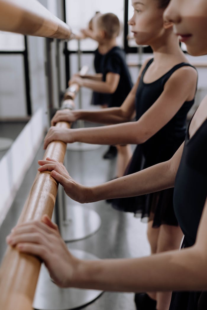 Young girls in black ballet attire practicing at a barre in a dance studio.